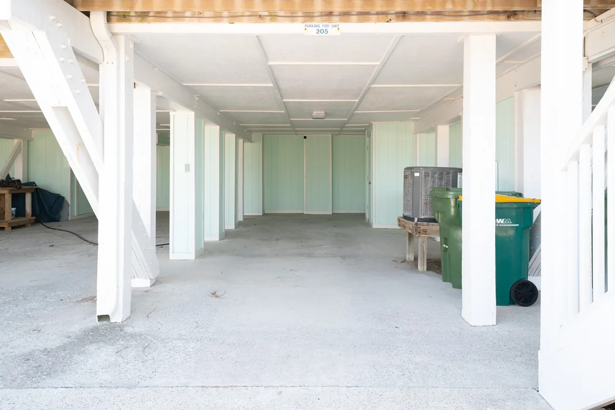 An open interior space with green and yellow trash bins and white support beams, likely a lower-level or garage area. The utilitarian mood hints at practical uses, such as storage or assigned parking.