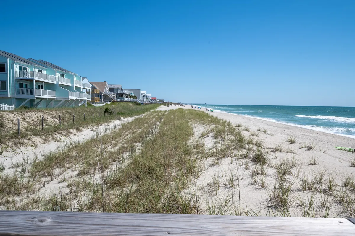 This image captures a classic stretch of beachfront with gentle ocean waves, a grassy dune, and a row of beachfront houses. Children and families can be imagined enjoying sandcastle building, swimming, and beachcombing along the shore, set against a relaxed coastal backdrop.