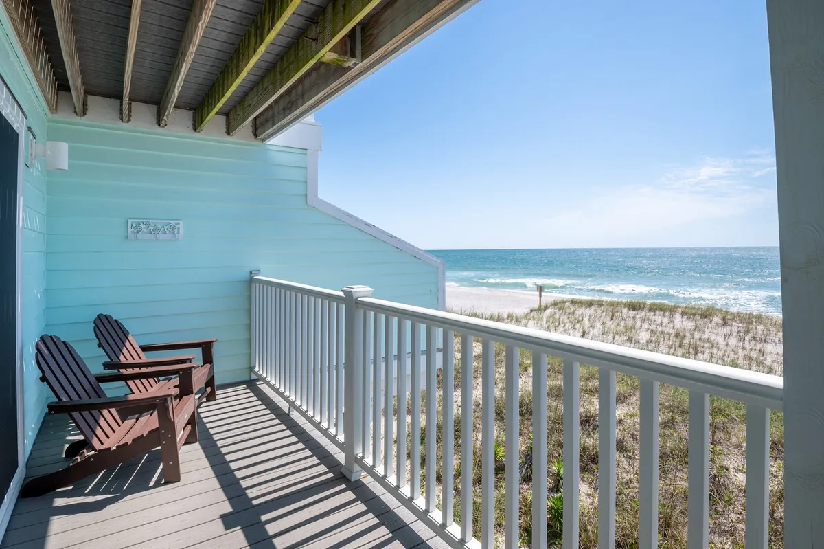 A serene exterior shot showing beach-facing deck chairs framed by a railing, with the expansive ocean and soft waves visible beyond. The lighting suggests either late afternoon or early morning, perfect for sunrise coffee or sunset cocktails. The empty chairs hint at privacy and relaxation, inviting couples or groups to unwind together while soaking up ocean views.