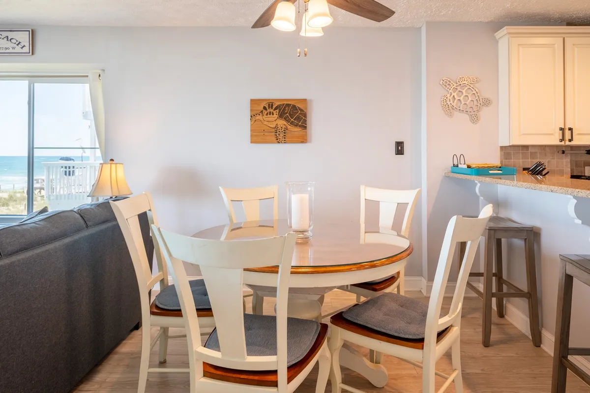 A cozy, open-plan dining and living area with a dining table set for four, a comfortable couch, and large windows that bring in ocean views. The decorative wall art adds a homey touch, and the layout fosters easy conversation and shared meals.