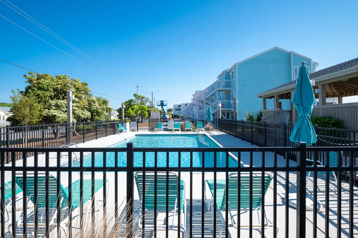 An exterior pool area view featuring a sparkling swimming pool surrounded by lounge chairs and umbrellas, with lush trees and a privacy fence creating a tranquil, resort-like atmosphere. The sun is low, suggesting the opportunity for sunset swims or leisurely afternoons by the pool.