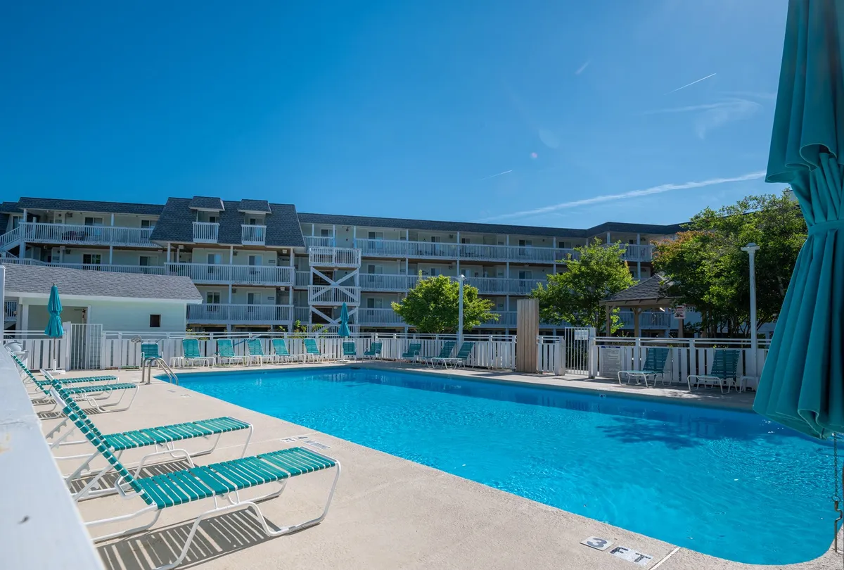 An inviting view of the resort's exterior pool area, with swimming pool, lounge chairs, and cheerful greenery surrounding the building facade. The atmosphere is lively yet relaxing, signaling opportunities for sunbathing, swimming, and group activities. The well-maintained communal space highlights the accessible amenities available to guests.