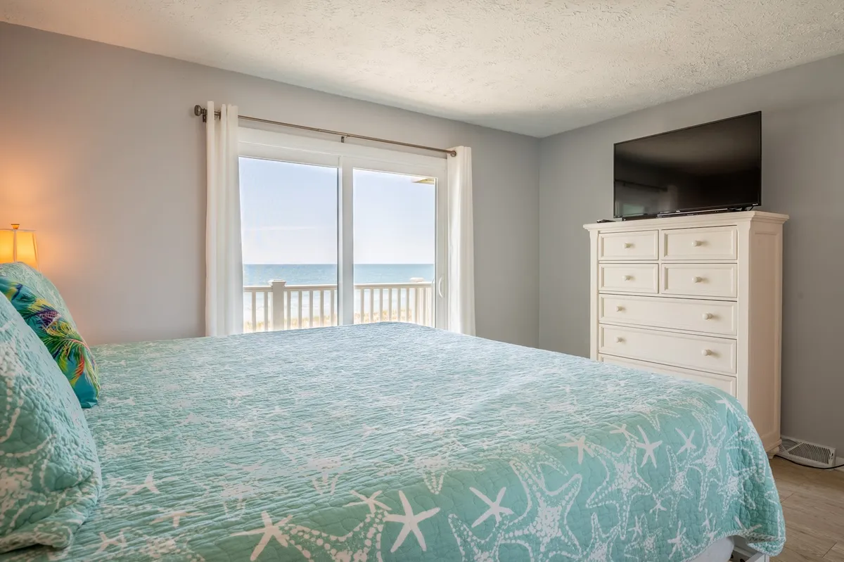 A tranquil bedroom with starfish-patterned bedding, a large window showcasing a stunning ocean view, and a TV placed on a dresser. The inviting space is perfect for relaxation and enjoying quiet moments by the shore.
