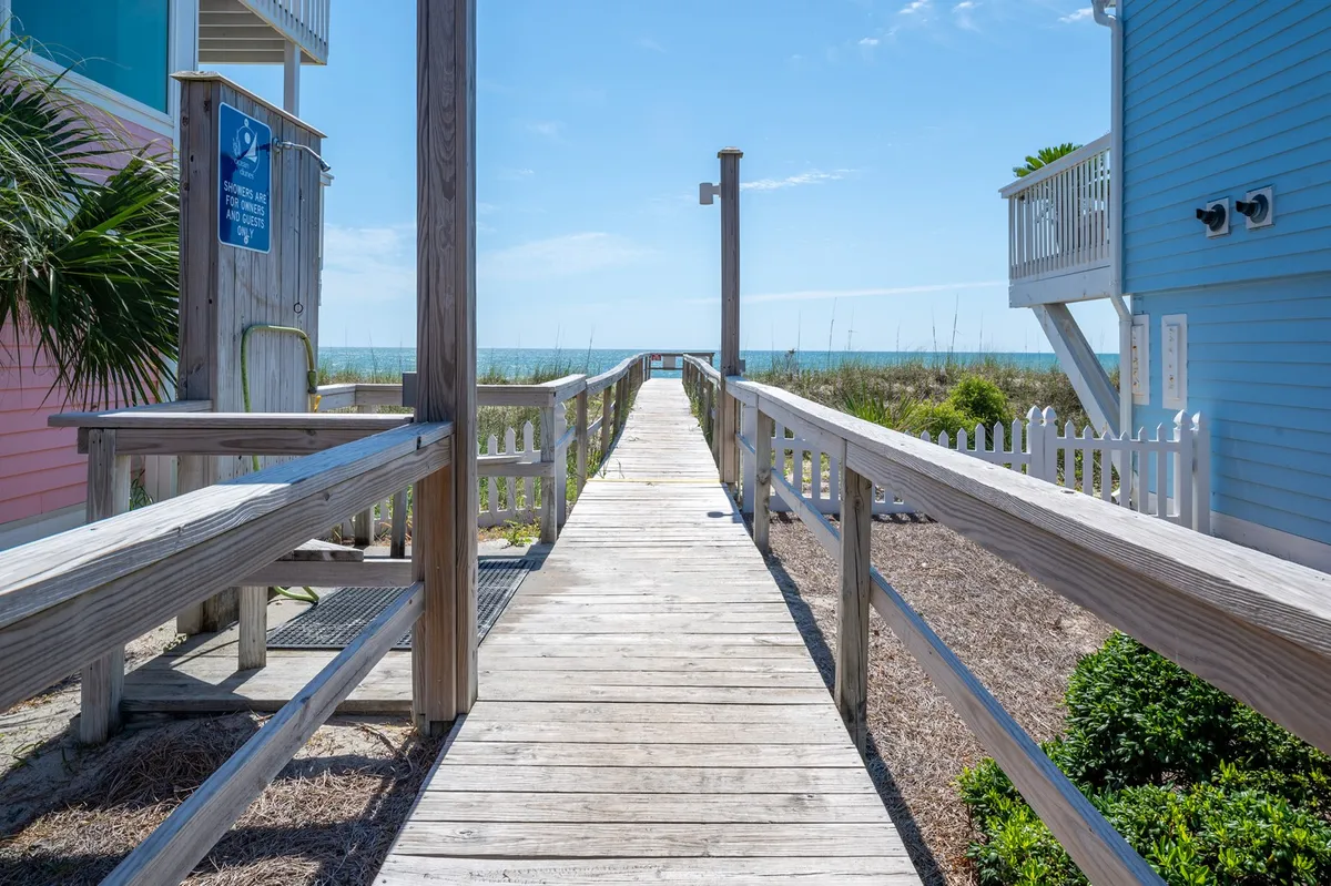 A wooden walkway leads through coastal greenery towards an expansive ocean view, evoking the feel of direct access to the beach. The tranquil setting hints at sunrise walks, picnics, and evening gatherings, with the sea as a constant backdrop.