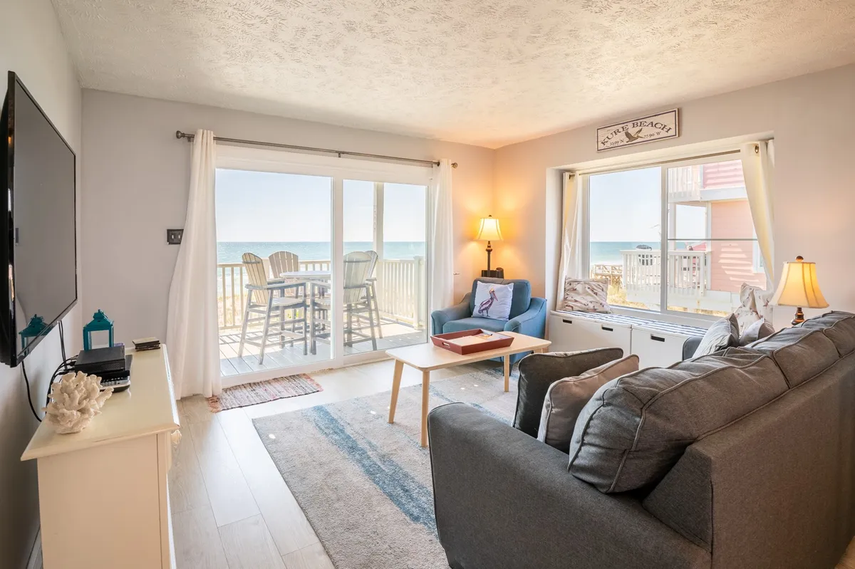 A bright, inviting living room featuring a comfortable sofa, coffee table with decorative accents, and large sliding glass doors that frame expansive ocean views. Warm lamps add to the cheerful atmosphere, and the space feels open and airy, perfect for relaxing or hosting movie nights.