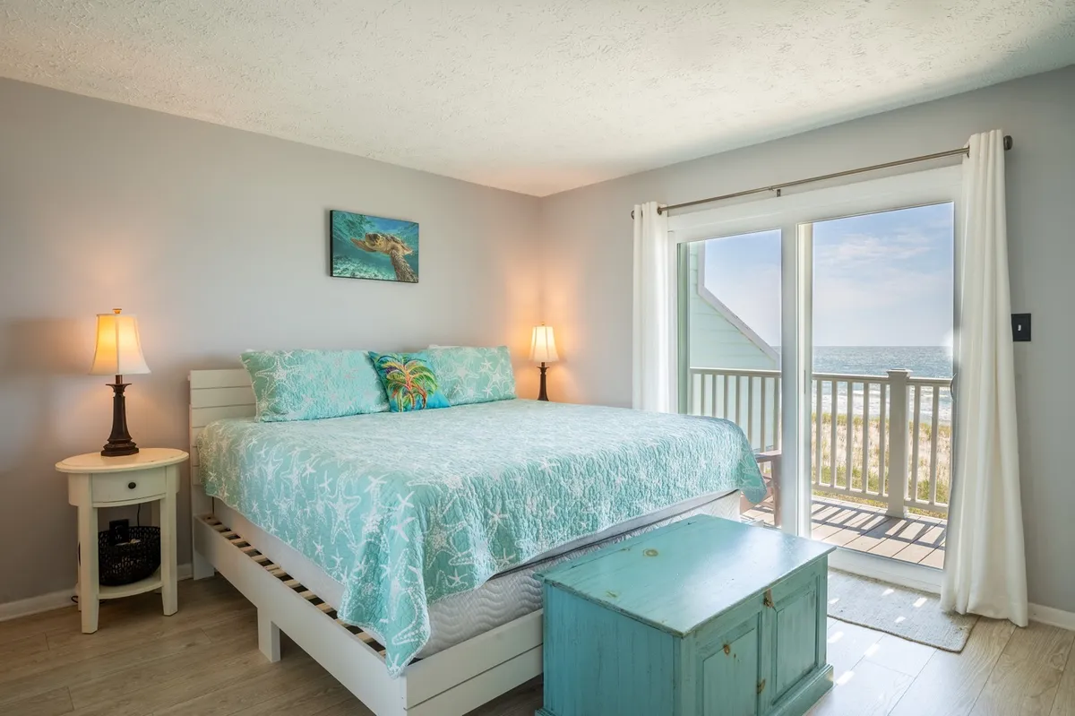 A serene primary bedroom featuring a bed with turquoise bedding, a bedside table with a lamp, a large window framing a sweeping ocean view, and painted artwork of sea life. Soft natural light fills the room, enhancing the calming coastal ambiance.