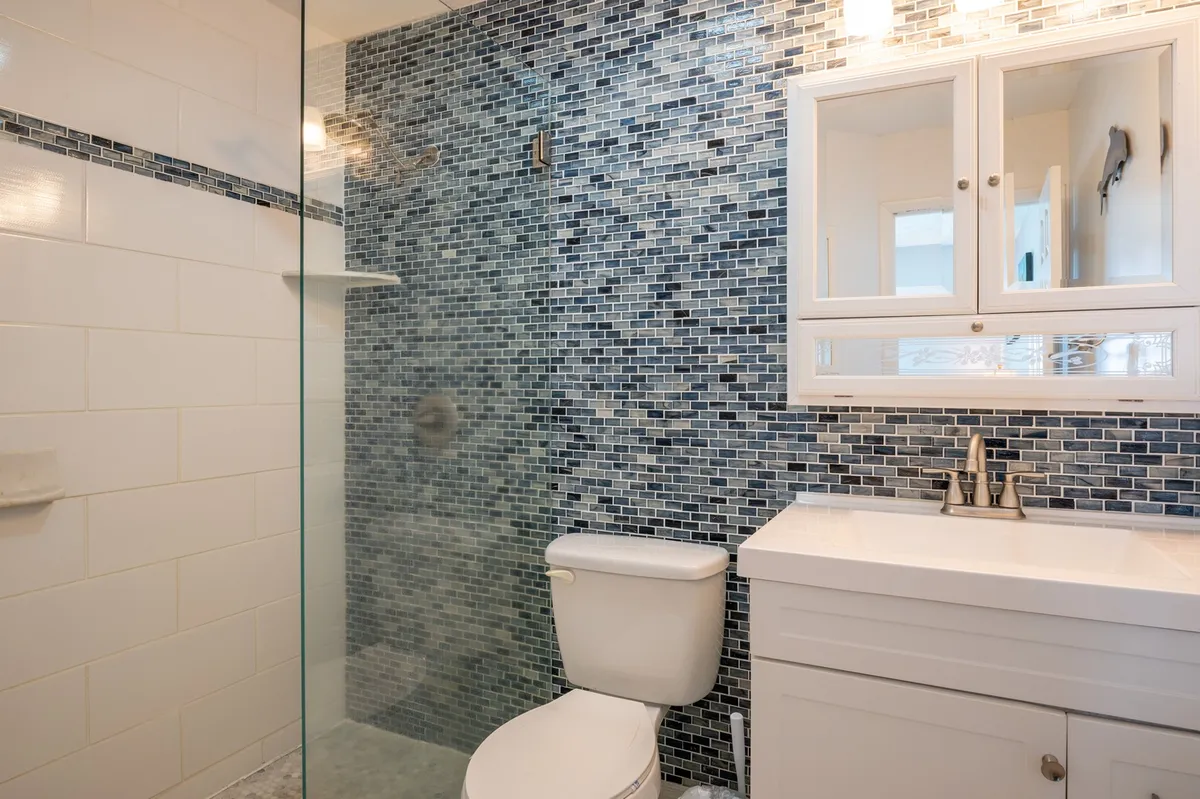 A modern bathroom with a striking mosaic tile shower wall, a white vanity topped with a quartz sink, and a mirrored cabinet above. The space feels refreshing and elegant, ideal for unwinding after sun-soaked days.