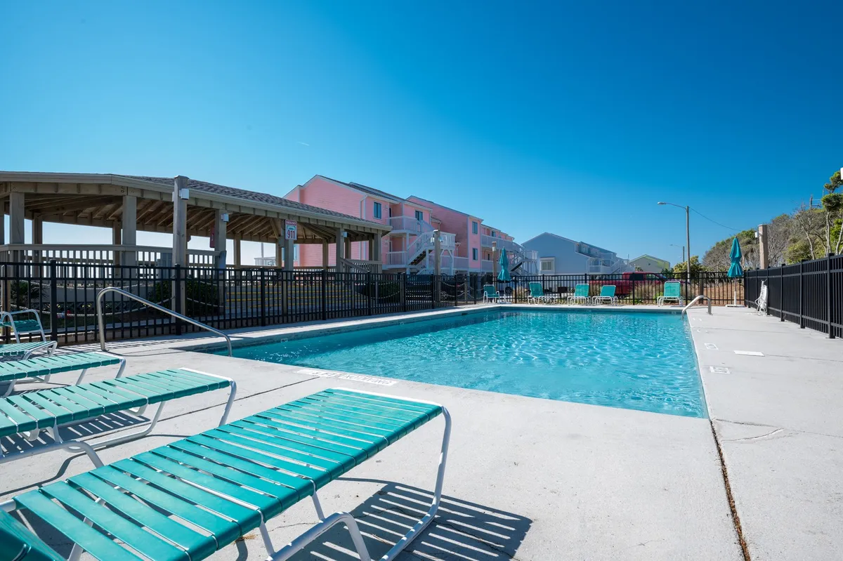 A vibrant, inviting outdoor swimming pool is surrounded by lounge chairs and colorful resort buildings, suggesting a lively, social atmosphere. The scene evokes memories of pool parties, sunbathing, and family relaxation under the sun.