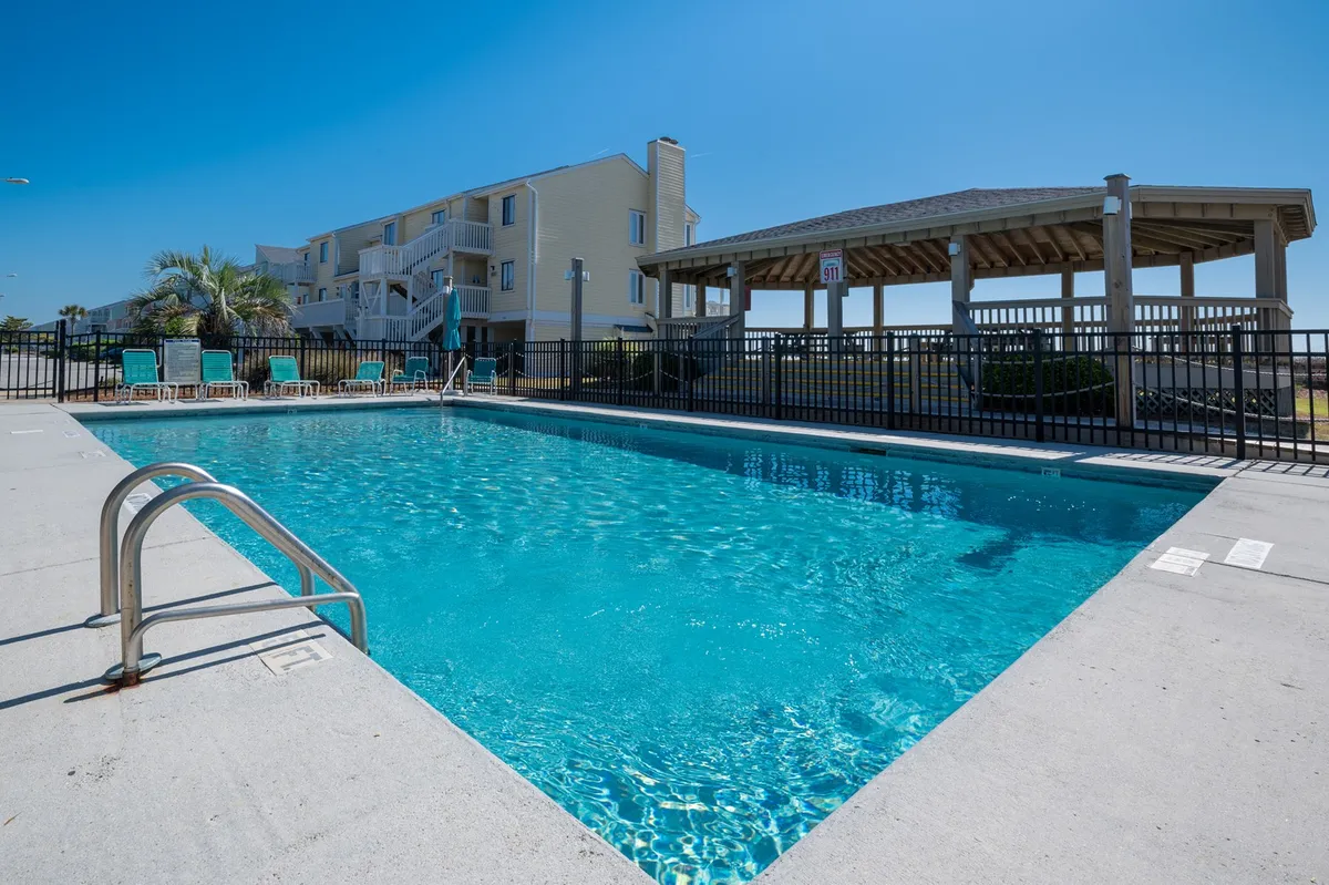 This image captures a vibrant outdoor swimming pool surrounded by seating areas, a gazebo, and lush palm trees. The scene is lively yet relaxing, with people enjoying swimming, sunbathing, and socializing under the gazebo, conveying a quintessential resort atmosphere.