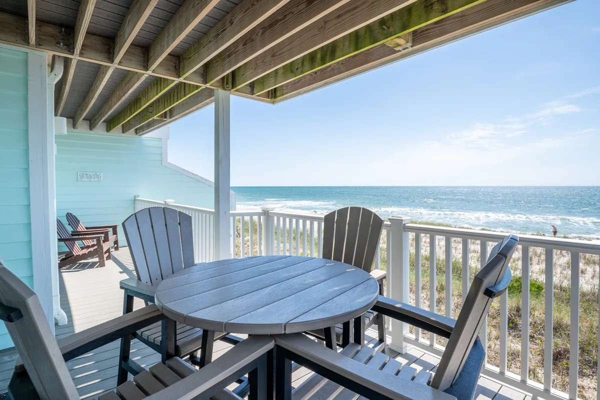 A spacious oceanfront deck with a wooden table and Adirondack chairs, overlooking the sparkling sea. The scene invites relaxing meals, sunset drinks, or simply enjoying the fresh sea breeze with loved ones.