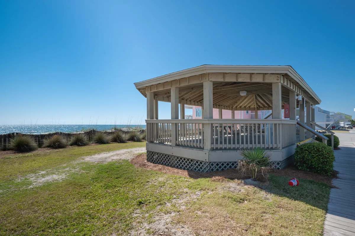 This exterior image features a gazebo set against an ocean view, bordered by a landscaped grass area. The scene illustrates a serene, light-hearted gathering space where guests can relax, socialize, or enjoy a picnic while children play on the lawn. The fresh sea breeze and open sky create a sense of connection to the outdoors and local environment.