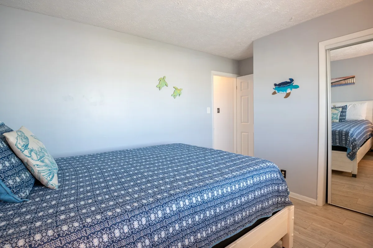 A peaceful bedroom scene with a bed made up in fresh blue patterned linens, colorful fish wall decals, and a mirror reflecting the room. The space feels clean and ready for new guests.