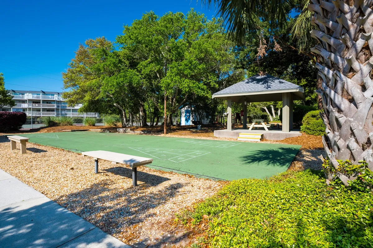 This exterior image centers on a green playing surface, a gazebo, benches, and surrounding trees. The area is designed for outdoor games like bocce or shuffleboard, as well as quiet moments in the gazebo or social events under the shade of trees. It exudes a welcoming, cheerful mood that invites group interaction.